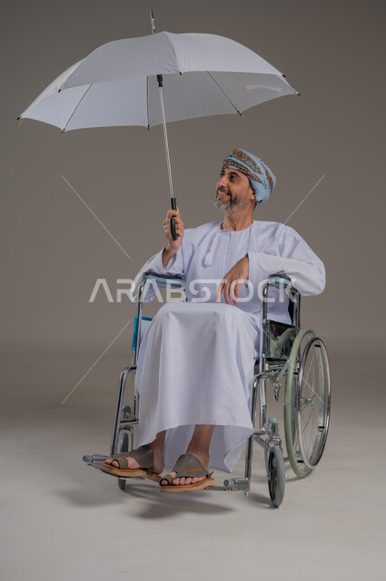 الحماية من الأمطار في الشتاء، دمج ذوي الهمم بالمجتمع العماني، التظلل من أشعة الشمس، بورتريه لرجل عربي خليجي عماني مقعد يرتدي الدشاشة والعمامة يجلس على كرسي متحرك يحمل بيده مظلة، صورة لكامل الجسم، خلفية رمادية