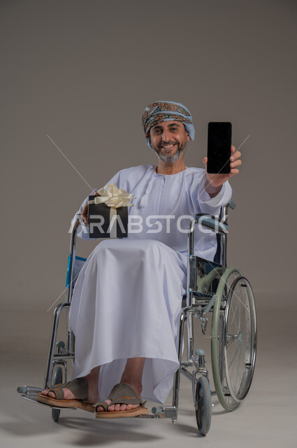 ذوي الإحتياجات الخاصة والهمم, مفهوم الاحتفال والهداية, بورتريه لشاب عربي خليجي عماني يرتدي العمامة والدشداشة يجلس على كرسي متحرك ويحمل بيده هدية وجهاز محمول بشاشة سوداء, صورة الجسم باطول الكامل،خلفية رمادية