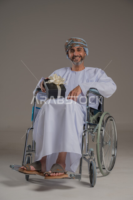 People with special needs and determination, the concept of celebration and guidance, a portrait of a young Arab Gulf Omani man wearing a turban and a dishdasha sitting in a wheelchair and holding a gift in his hand, a full-length body image, gray background