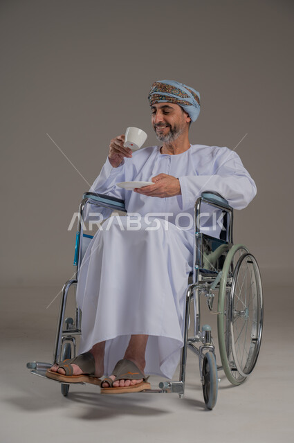 التمسك بالقيم الكريمة المتوارثة من الأجداد، بورتريه لرجل عربي خليجي عماني من ذوي الاحتياجات الخاصة يرتدي العمامة والدشداشة يجلس على كرسي متحرك يحمل بيده فنجان من القهوة، الاستمتاع بالمشروبات الساخنة، صورة الجسم بالطول الكامل، خلفية رمادية