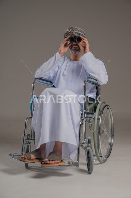 استعمال المنظار للكشف عن الاشياء البعيدة، ذوي الإحتياجات الخاصة والهمم, بورتريه لشاب عربي خليجي عماني من ذوي الاحتياجات الخاصة يرتدي العمامة والدشداشة يجلس على كرسي متحرك، خلفية رمادية