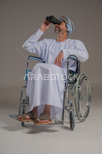 استعمال المنظار للكشف عن الاشياء البعيدة، ذوي الإحتياجات الخاصة والهمم, بورتريه لشاب عربي خليجي عماني من ذوي الاحتياجات الخاصة يرتدي العمامة والدشداشة يجلس على كرسي متحرك، خلفية رمادية