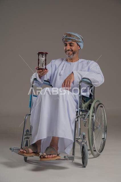 مفهوم إدارة الأعمال وتنظيم الجدول الزمني، تحديد مواعيد وأوقات العمل، القيام بمراقبة توقيت الرمل, بورتريه لرجل عماني خليجي من ذوي الاحتياجات الخاصة  يرتدي الزي الدشاشة والعمامة يحمل بيده الساعة الرملية الزجاجية (البنكام)، خلفية رمادية