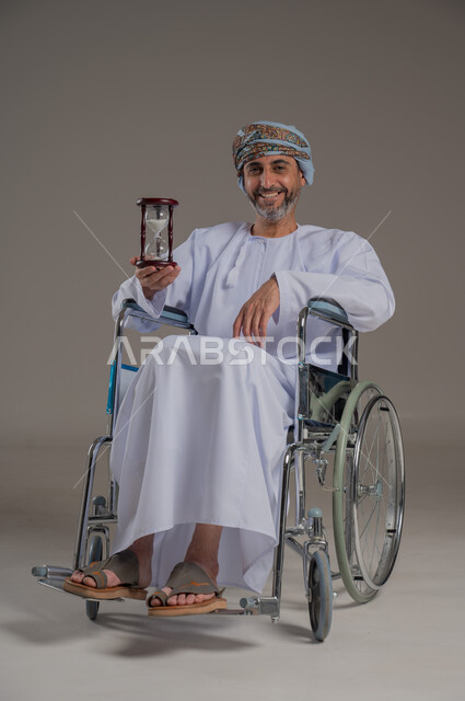 مفهوم إدارة الأعمال وتنظيم الجدول الزمني، تحديد مواعيد وأوقات العمل، القيام بمراقبة توقيت الرمل, بورتريه لرجل عماني خليجي من ذوي الاحتياجات الخاصة  يرتدي الزي الدشاشة والعمامة يحمل بيده الساعة الرملية الزجاجية (البنكام)، خلفية رمادية