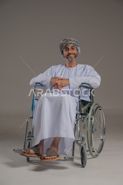 استخدام كراسي المعاقين للمساعده على الحركه والتنقل، اسلوب حياة ذوي الهمم، تحسين جودة حياة ذوي الاعاقه الحركيه، بورتريه لرجل عربي خليجي عماني مقعد يرتدي الدشاشة والعمامة  يجلس على كرسي متحرك، صورة لكامل الجسم، خلفية رمادية