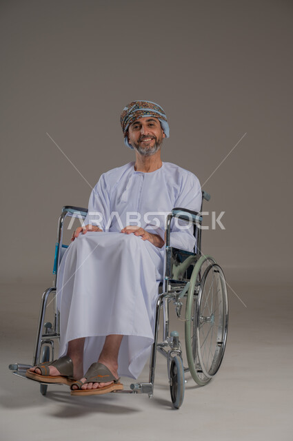 استخدام كراسي المعاقين للمساعده على الحركه والتنقل، اسلوب حياة ذوي الهمم، تحسين جودة حياة ذوي الاعاقه الحركيه، بورتريه لرجل عربي خليجي عماني مقعد يرتدي الدشاشة والعمامة  يجلس على كرسي متحرك، صورة لكامل الجسم، خلفية رمادية