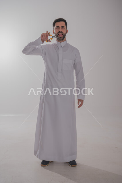 عدم القدرة على التوقف من الكلام، الاهتمام بالمظهر الخارجي والأناقة، مفهوم السكوت والصمت، بورتريه لرجل عربي خليجي سعودي يرتدي الثوب التقليدي يحمل بيده مقص ينظر الى الكاميرا بتعابير الجدية، الوقوف باستقامة وثبات، صورة الجسم بالطول الكامل، خلفية رمادية