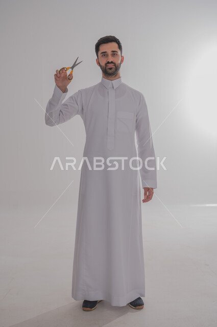 عدم القدرة على التوقف من الكلام، الاهتمام بالمظهر الخارجي والأناقة، مفهوم السكوت والصمت، بورتريه لرجل عربي خليجي سعودي يرتدي الثوب التقليدي يحمل بيده مقص ينظر الى الكاميرا بتعابير الجدية، الوقوف باستقامة وثبات، صورة الجسم بالطول الكامل، خلفية رمادية