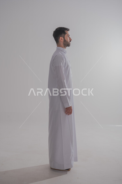 Concern about appearance and elegance, concept of masculinity and self-confidence, side portrait of a Saudi Arabian Gulf man wearing traditional thobe looking at something with an expression of pleasure, standing straight and steady, full-length body image, gray background