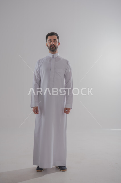 مفهوم الرجولة والثقة بالنفس، الوقوف باستعداد واستقامة، الاهتمام بالمظهر الخارجي والاناقة، بورتريه لرجل عربي خليجي سعودي يرتدي الثوب الأبيض يقف باستقامة وينظر الى الكاميرا بتعابير الجدية والثقة بالنفس، صورة الجسم بالطول الكامل، خلفية رمادية