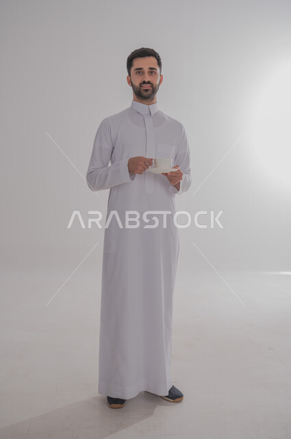 الاستمتاع بالمشروبات الساخنة، مفهوم الرجولة والثقة بالنفس، بورتريه لرجل عربي خليجي سعودي يرتدي الثوب التقليدي يحمل بيده كوب من القهوة ينظر الى الكاميرا بتعابير السرور، الوقوف باستقامة وثبات، صورة الجسم بالطول الكامل، خلفية رمادية