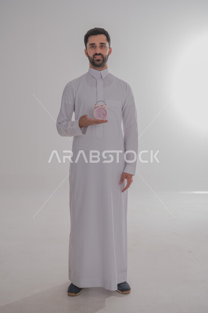 Timer, concept of masculinity and self-confidence, portrait of a Saudi Arabian Gulf man wearing traditional thobe holding a small watch looking at the camera with an expression of pleasure, standing straight and steady, full body image, gray background