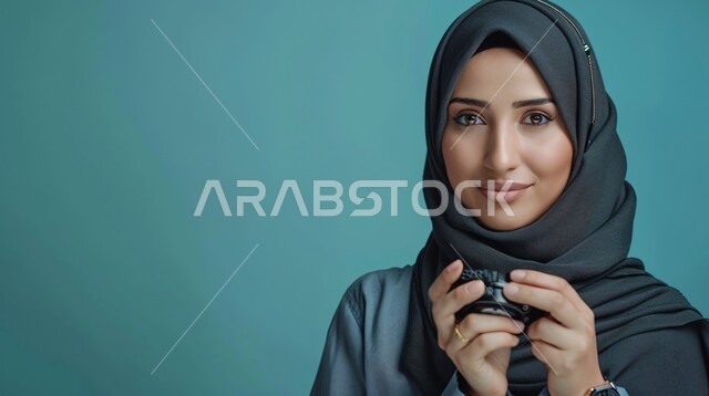 مفهوم الأنوثة والنعومة، الاهتمام بالأناقة والمظهر الخارجي، بورتريه مقرب لإمرأة عربية خليجية سعودية مبتسمة ترتدي حجاب وعباءة  تنظر الى الكاميرا بتعابير السرور وتحمل بيدها كوب من القهوة، إيماءات الثقة بالنفس, خلفية زرقاء