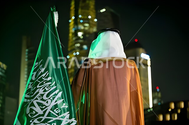 Celebrating Flag Day, March 11, remembering history and national identity, commemorating the Saudi National Day, September 23, a picture from behind of a Saudi Gulf Arab man wearing the Saudi dress holding the national flag in his hand, Saudi National Day, background of the King Abdullah Towers