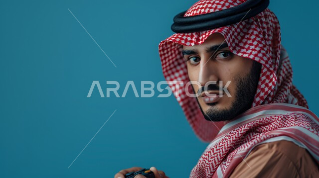 قضاء أوقات مسلية باللعب في الألعاب الالكترونية الترفيهيه، بورتريه مقرب لشاب عربي خليجي سعودي يرتدي الثوب التقليدي والشماغ يجلس يمسك بيده عصا التحكم، المتعة والتسلية بالفيديو جيمز، النظر الى الكاميرا بتعابير الاندماج، خلفية زرقاء