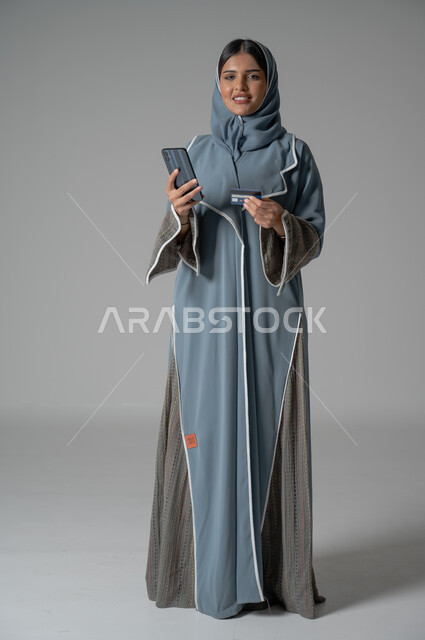 الأهتمام بالمظهر الخارجي, بورتريه لإمرأة عربية خليجية سعودية محجبة ترتدي عباءة تحمل الهاتف المحمول بايماءات السعادة والسرور، صورة الجسم بالطول الكامل, خلفية رمادية