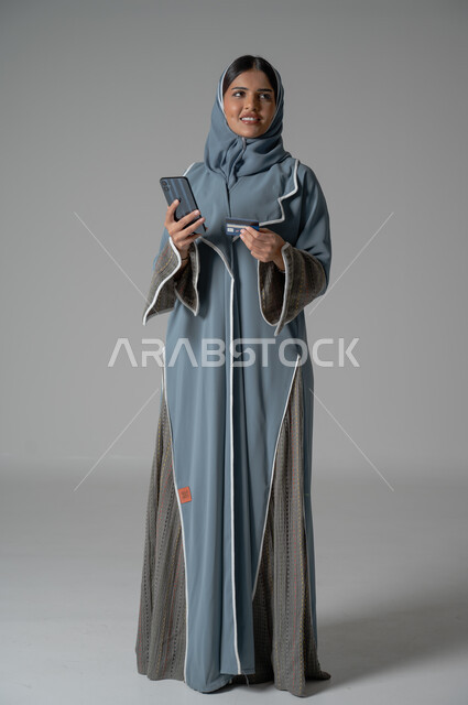 الأهتمام بالمظهر الخارجي, بورتريه لإمرأة عربية خليجية سعودية محجبة ترتدي عباءة تحمل الهاتف المحمول بايماءات السعادة والسرور، صورة الجسم بالطول الكامل, خلفية رمادية