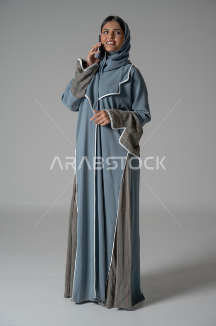 الأهتمام بالمظهر الخارجي, بورتريه لإمرأة عربية خليجية سعودية محجبة ترتدي عباءة تقوم باجراء مكالمة هاتفية عبر الجوال بايماءات السعادة والسرور، صورة الجسم بالطول الكامل, خلفية رمادية