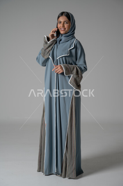 الأهتمام بالمظهر الخارجي, بورتريه لإمرأة عربية خليجية سعودية محجبة ترتدي عباءة تقوم باجراء مكالمة هاتفية عبر الجوال بايماءات السعادة والسرور، صورة الجسم بالطول الكامل, خلفية رمادية