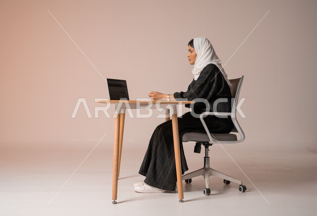 إدارة شؤون الأعمال وتنظيمه, احتراف وظائف الحاسب والتكنولوجيا، بورتريه لإمراه عربيه سعوديه خليجيه محجبة ترتدي عباءة سوداء  تجلس على كرسي مريح تنظر الى جهاز الحاسوب بايماءات الانزعاج، إنجاز المهام المكتبية، خلفية بيج