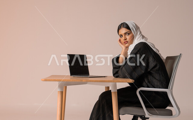 Business Administration and Organization, Professional Computer and Technology Jobs, Portrait of a Saudi Arabian Gulf Woman in a Veil Wearing a Black Abaya Sitting on a Comfortable Chair Looking at the Camera with Annoyed Gestures, Completing Office Tasks, Beige Background