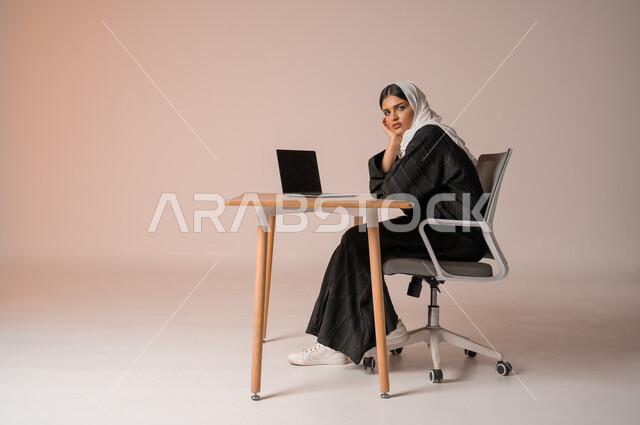 Business Administration and Organization, Professional Computer and Technology Jobs, Portrait of a Saudi Arabian Gulf Woman in a Veil Wearing a Black Abaya Sitting on a Comfortable Chair Looking at the Camera with Annoyed Gestures, Completing Office Tasks, Beige Background