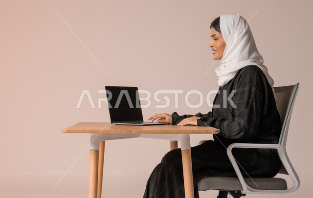 استخدام التقنيات الحديثة والمتطورة، بورتريه لامراة عربية خليجية سعودية محجبة مبتسمة ترتدي العباءة السوداء تجلس على الكرسي أمام الحاسوب المحمول بايماءات السعادة والسرور، التواصل مع الأهل و الأصدقاء عن بعد، إدارة شؤون الأعمال وتنظيمه، خلفية بيج