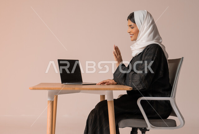 اجتماعات عمل أونلاين, القاء التحية والسلام عن بعد، مكالمات فيديو وبث مباشر, بورتريه لامرأة عربية خليجية سعودية مبتسمة محجبة ترتدي العباءة تجلس على الكرسي ترفع يدها للتلويح وتقوم باستخدام جهاز الحاسوب, التواصل مع الأهل والأصدقاء, خلفية بيج