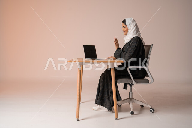 اجتماعات عمل أونلاين, القاء التحية والسلام عن بعد، مكالمات فيديو وبث مباشر, بورتريه لامرأة عربية خليجية سعودية مبتسمة محجبة ترتدي العباءة تجلس على الكرسي ترفع يدها للتلويح وتقوم باستخدام جهاز الحاسوب, التواصل مع الأهل والأصدقاء, خلفية بيج