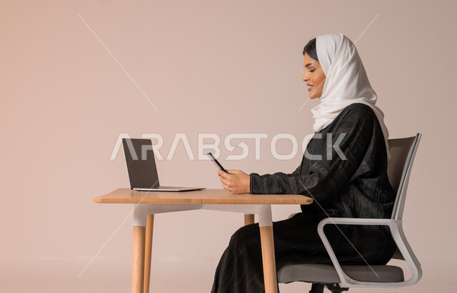 Using modern and advanced technologies, portrait of a smiling veiled Saudi Arabian Gulf woman wearing a black abaya sitting on a chair in front of a laptop and holding a mobile phone in her hand with gestures of happiness and pleasure, communicating with family and friends remotely, managing and organizing business affairs, beige background