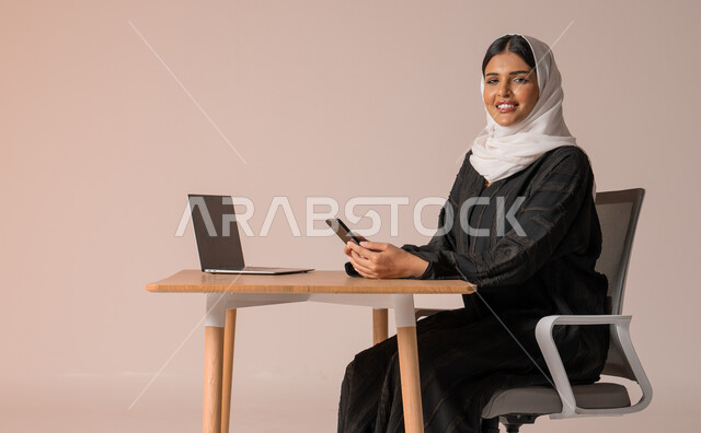 استخدام التقنيات الحديثة والمتطورة، بورتريه لامراة عربية خليجية سعودية محجبة مبتسمة ترتدي العباءة السوداء تجلس على الكرسي أمام الحاسوب المحمول وتمسك بيدها الجوال، التواصل مع الأهل و الأصدقاء عن بعد، إدارة شؤون الأعمال وتنظيمه، النظر الى الكاميرا بايماءات السعادة والسرور، خلفية بيج