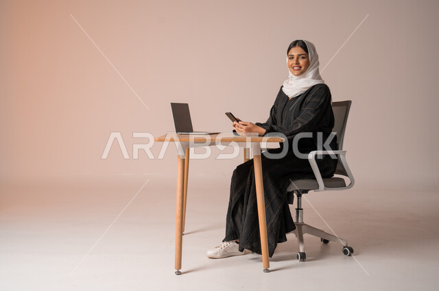 استخدام التقنيات الحديثة والمتطورة، بورتريه لامراة عربية خليجية سعودية محجبة مبتسمة ترتدي العباءة السوداء تجلس على الكرسي أمام الحاسوب المحمول وتمسك بيدها الجوال، التواصل مع الأهل و الأصدقاء عن بعد، إدارة شؤون الأعمال وتنظيمه، النظر الى الكاميرا بايماءات السعادة والسرور، خلفية بيج