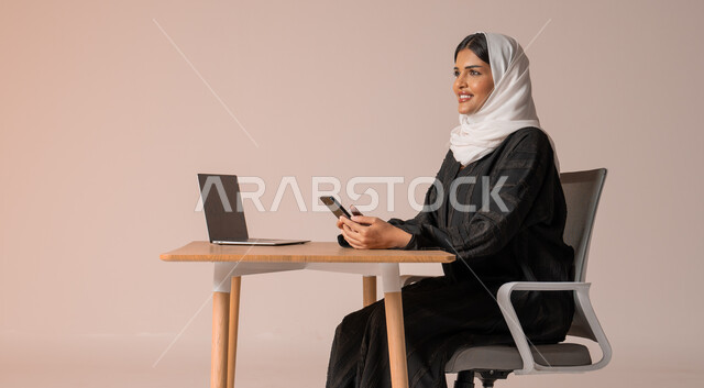 استخدام التقنيات الحديثة والمتطورة، بورتريه لامراة عربية خليجية سعودية محجبة مبتسمة ترتدي العباءة السوداء تجلس على الكرسي أمام الحاسوب المحمول وتمسك بيدها الجوال بايماءات السعادة والسرور، التواصل مع الأهل و الأصدقاء عن بعد، إدارة شؤون الأعمال وتنظيمه,  خلفية بيج