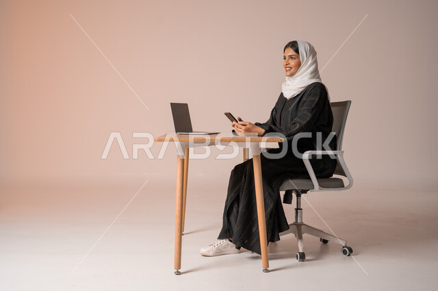 استخدام التقنيات الحديثة والمتطورة، بورتريه لامراة عربية خليجية سعودية محجبة مبتسمة ترتدي العباءة السوداء تجلس على الكرسي أمام الحاسوب المحمول وتمسك بيدها الجوال بايماءات السعادة والسرور، التواصل مع الأهل و الأصدقاء عن بعد، إدارة شؤون الأعمال وتنظيمه,  خلفية بيج