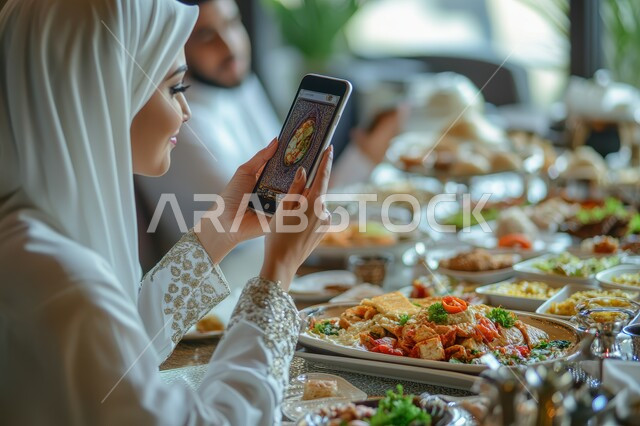 أسرة عربية خليجية سعودية تجلس في المطعم، مائدة مليئة بالمأكولات والمشروبات المختلفة، إمرأة محجبة تقوم بالتقاط صور تذكارية لسفرة الطعام باستخدام الهاتف المحمول، قضاء أوقات ممتعة في الخارج، عطلة نهاية الاسبوع، أجواء عائلية سعيدة