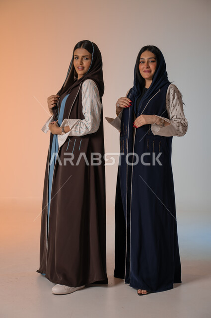 Wearing the Gulf abaya and sunglasses, gestures of confidence and standing in front of the camera, professionalism in displaying the latest fashion and beauty trends, a portrait of two Saudi Gulf Arab models, a full-length body photo, Saudi women’s products, buying fashion and accessories, a colored background.