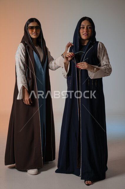Wearing the Gulf abaya and sunglasses, gestures of confidence and standing in front of the camera, professionalism in displaying the latest fashion and beauty trends, a portrait of two Saudi Gulf Arab models, a full-length body photo, Saudi women’s products, buying fashion and accessories, a colored background.