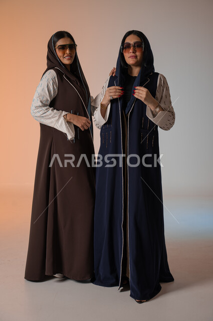 Wearing the Gulf abaya and sunglasses, gestures of confidence and standing in front of the camera, professionalism in displaying the latest fashion and beauty trends, a portrait of two Saudi Gulf Arab models, a full-length body photo, Saudi women’s products, buying fashion and accessories, a colored background.