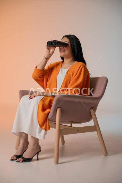 التخطيط للمستقبل وتحديد الأهداف والطموحات، بورتريه لامرأة عربية خليجية سعودية أنيقة ترتدي الزي الكاجوال تجلس على كرسي تحمل بيدها منظار ثنائي العينين وتنظر فيه بإيماءات الحماس والسرور، استخدام الأدوات والأجهزة التقنية الحديثة لرؤية الأشياء البعيدة، خلفية ملونة