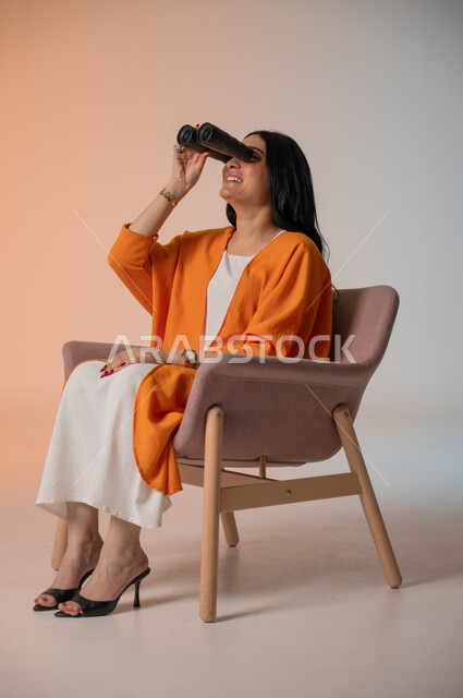 التخطيط للمستقبل وتحديد الأهداف والطموحات، بورتريه لامرأة عربية خليجية سعودية أنيقة ترتدي الزي الكاجوال تجلس على كرسي تحمل بيدها منظار ثنائي العينين وتنظر فيه بإيماءات الحماس والسرور، استخدام الأدوات والأجهزة التقنية الحديثة لرؤية الأشياء البعيدة، خلفية ملونة