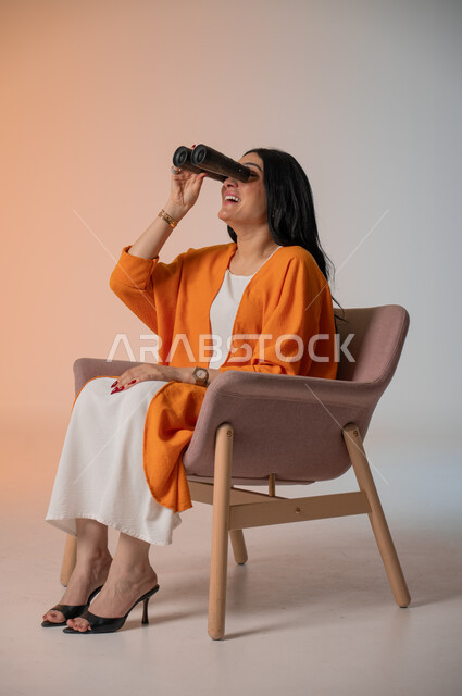 التخطيط للمستقبل وتحديد الأهداف والطموحات، بورتريه لامرأة عربية خليجية سعودية أنيقة ترتدي الزي الكاجوال تجلس على كرسي تحمل بيدها منظار ثنائي العينين وتنظر فيه بإيماءات الحماس والسرور، استخدام الأدوات والأجهزة التقنية الحديثة لرؤية الأشياء البعيدة، خلفية ملونة