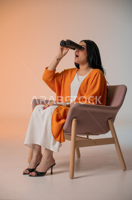 التخطيط للمستقبل وتحديد الأهداف والطموحات، بورتريه لامرأة عربية خليجية سعودية أنيقة ترتدي الزي الكاجوال تجلس على كرسي تحمل بيدها منظار ثنائي العينين وتنظر فيه بإيماءات الحماس والسرور، استخدام الأدوات والأجهزة التقنية الحديثة لرؤية الأشياء البعيدة، خلفية ملونة