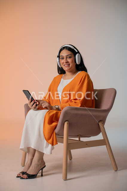 Listening to and enjoying your favorite music, having fun, browsing social media, portrait of a Saudi Arabian Gulf woman in casual wear sitting on a chair with headphones and holding a mobile phone, using advanced technology, colorful background