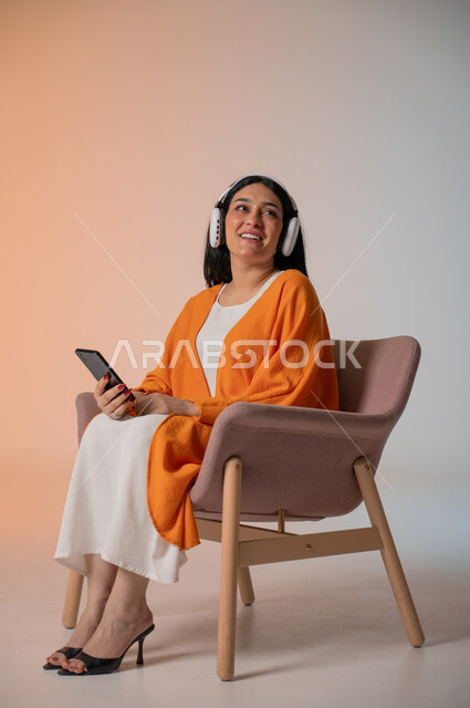الاستماع إلى الموسيقا المفضلة والاستمتاع بها، قضاء أوقات الفراغ بشكل ممتع، تصفح مواقع التواصل الاجتماعي، بورتريه لامرأة عربية خليجية سعودية ترتدي الزي الكاجوال تجلس على كرسي تضع الهيدفون وتحمل بيدها الهاتف المحمول، استخدام التكنولوجيا المتطورة ، خلفية ملونة