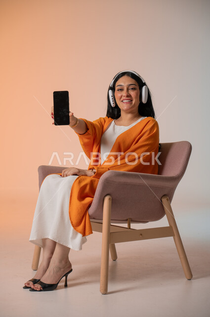عرض شاشة جوال سوداء فارغة، الاستماع إلى الموسيقا المفضلة والاستمتاع بها، تصفح مواقع التواصل الاجتماعي، بورتريه لامرأة عربية خليجية سعودية ترتدي الزي الكاجوال تجلس على كرسي تضع الهيدفون وتحمل بيدها الهاتف المحمول، استخدام التكنولوجيا المتطورة ، خلفية ملونة