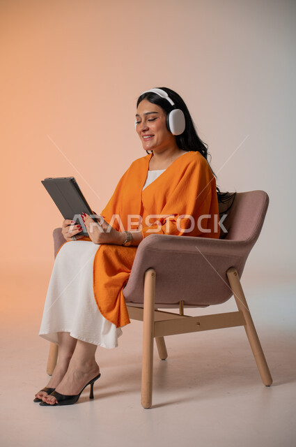 Using advanced technology and tablets to spend free time, listening to and enjoying favorite music, browsing social media, portrait of a Saudi Arabian Gulf woman wearing casual clothes sitting on a chair with headphones and holding a tablet in her hand, colorful background