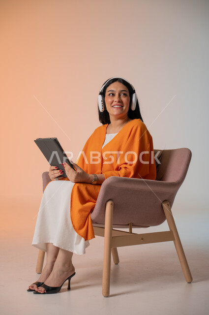 استخدام التكنولوجيا المتطورة والجهزة اللوحية في تمضية وقت الفراغ، الاستماع إلى الموسيقا المفضلة والاستمتاع بها، تصفح مواقع التواصل الاجتماعي، بورتريه لامرأة عربية خليجية سعودية ترتدي الزي الكاجوال تجلس على كرسي تضع الهيدفون وتحمل بيدها التابلت، خلفية ملونة