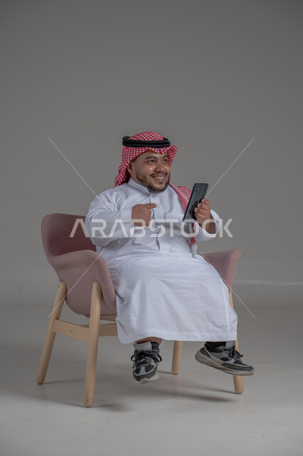قضاء وقت ممتع, بورتريه لرجل عربي خليجي سعودي قصير القامة يجلس على كرسي مريح ويستخدم الهاتف المحمول ، استخدام البرامج والتطبيقات الالكترونية، التسوق الالكتروني عبر الانترنت، تصفح المواقع الالكترونية والتواصل الاجتماعي، خلفية رمادية