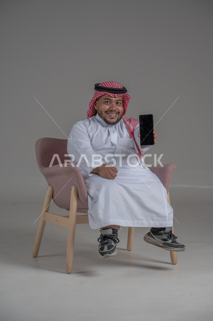 قضاء وقت ممتع, بورتريه لرجل عربي خليجي سعودي قصير القامة يجلس على كرسي مريح ويستخدم الهاتف المحمول ، استخدام البرامج والتطبيقات الالكترونية، التسوق الالكتروني عبر الانترنت، تصفح المواقع الالكترونية والتواصل الاجتماعي، خلفية رمادية