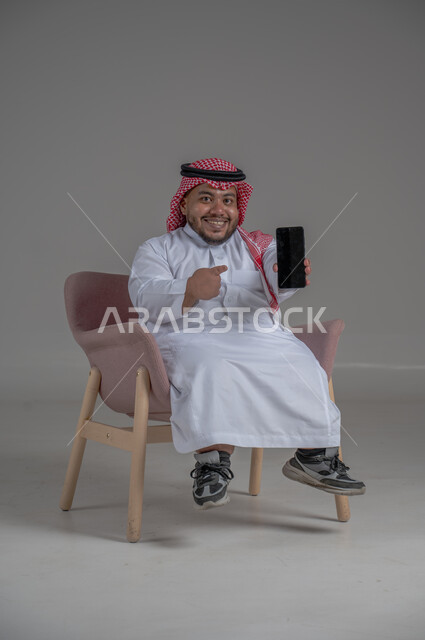 قضاء وقت ممتع, بورتريه لرجل عربي خليجي سعودي قصير القامة يجلس على كرسي مريح ويستخدم الهاتف المحمول ، استخدام البرامج والتطبيقات الالكترونية، التسوق الالكتروني عبر الانترنت، تصفح المواقع الالكترونية والتواصل الاجتماعي، خلفية رمادية